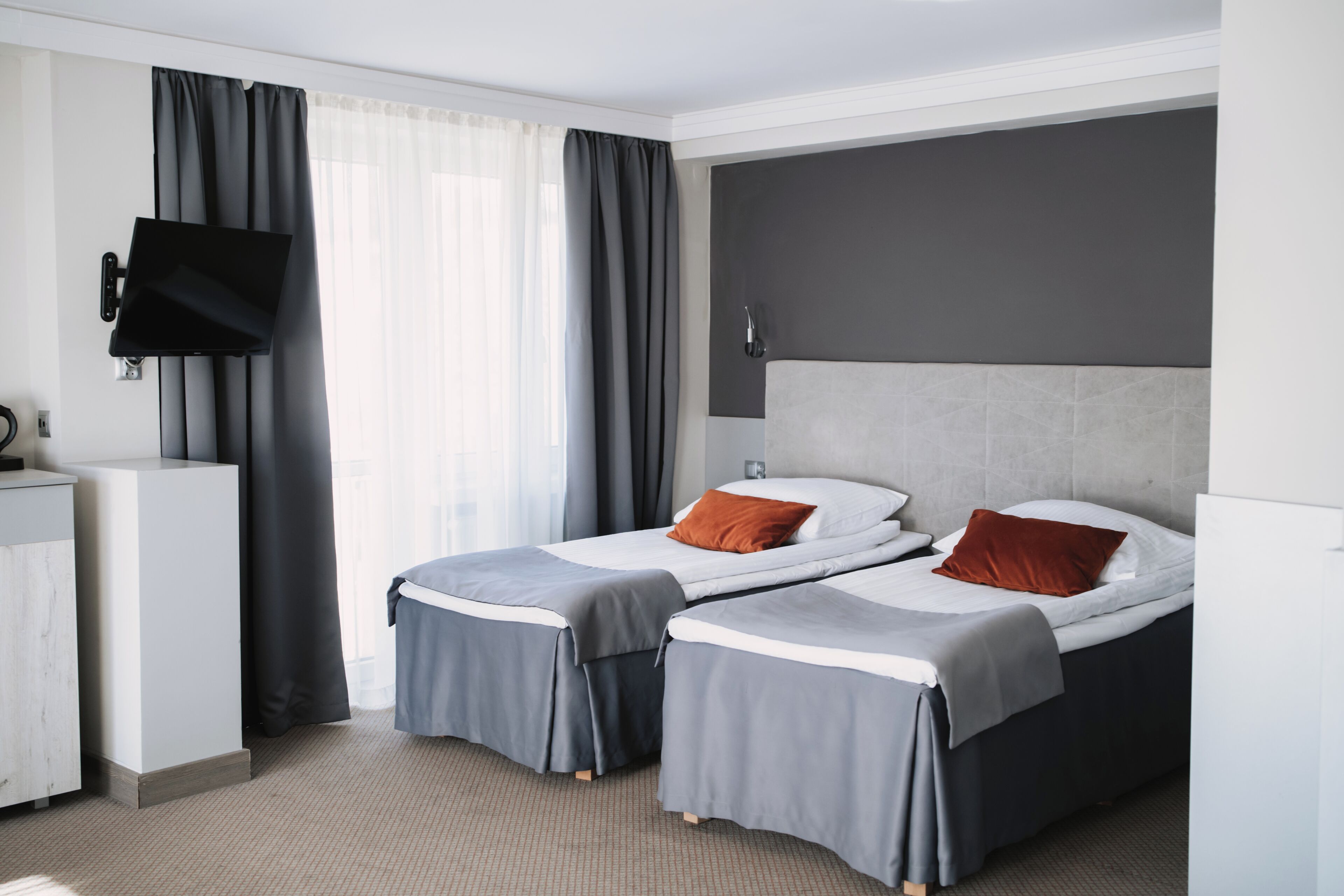 A hotel room with two beds, a desk, a flat-screen TV, and a large window with curtains.
