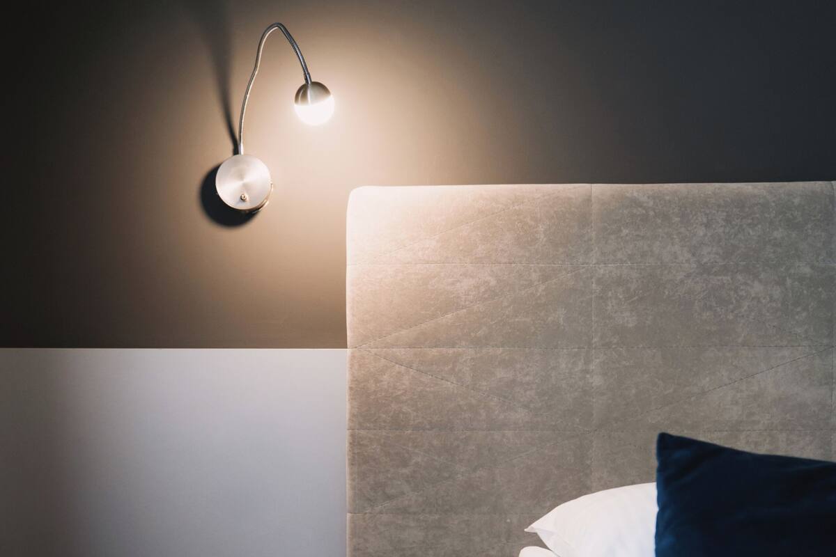 Una cama bien hecha con cabecera, una mesita de noche con cajón y una lámpara de pared.