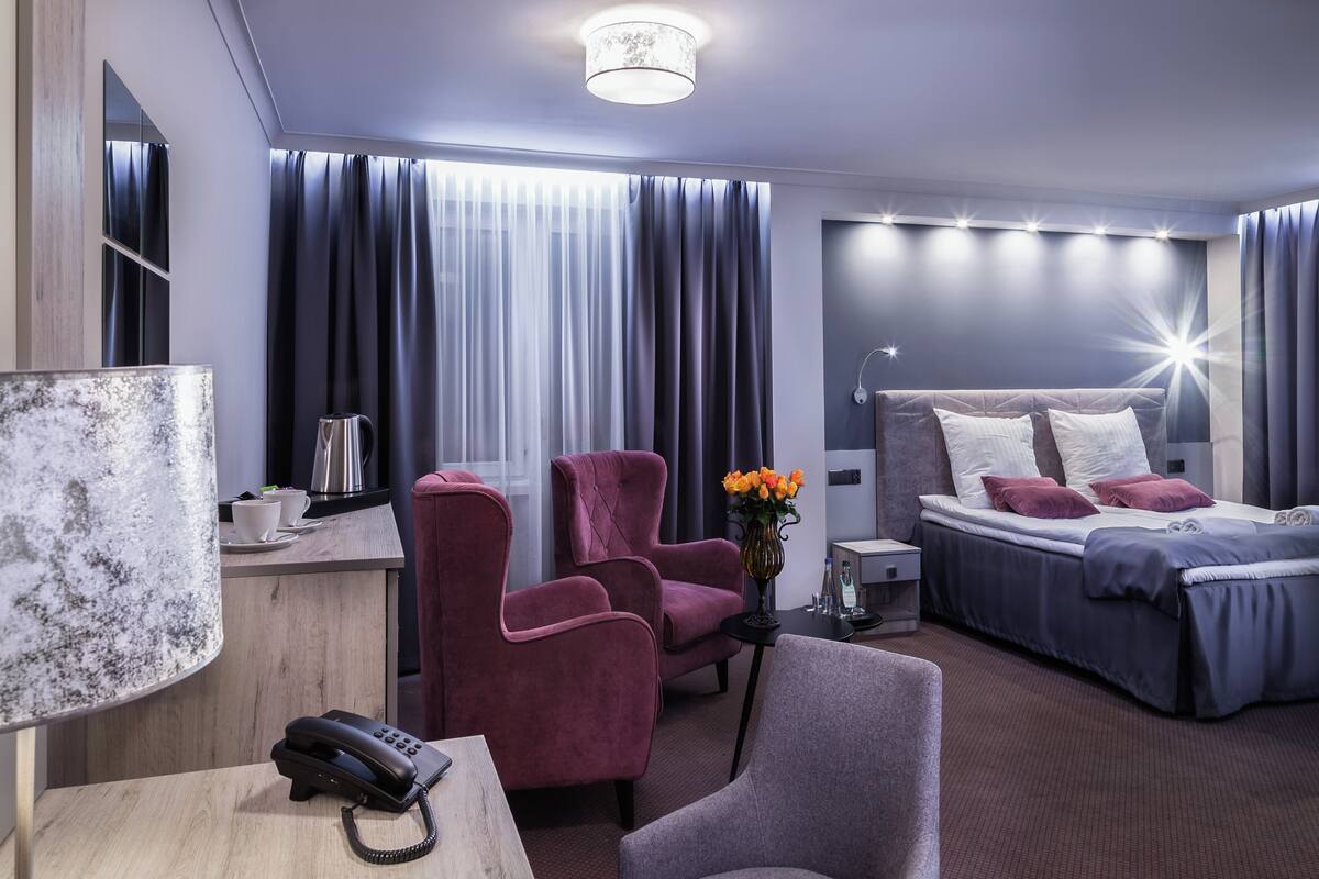 Habitación de hotel moderna con una cama, dos sillones, un escritorio con un teléfono, una cafetera y un jarrón con flores.