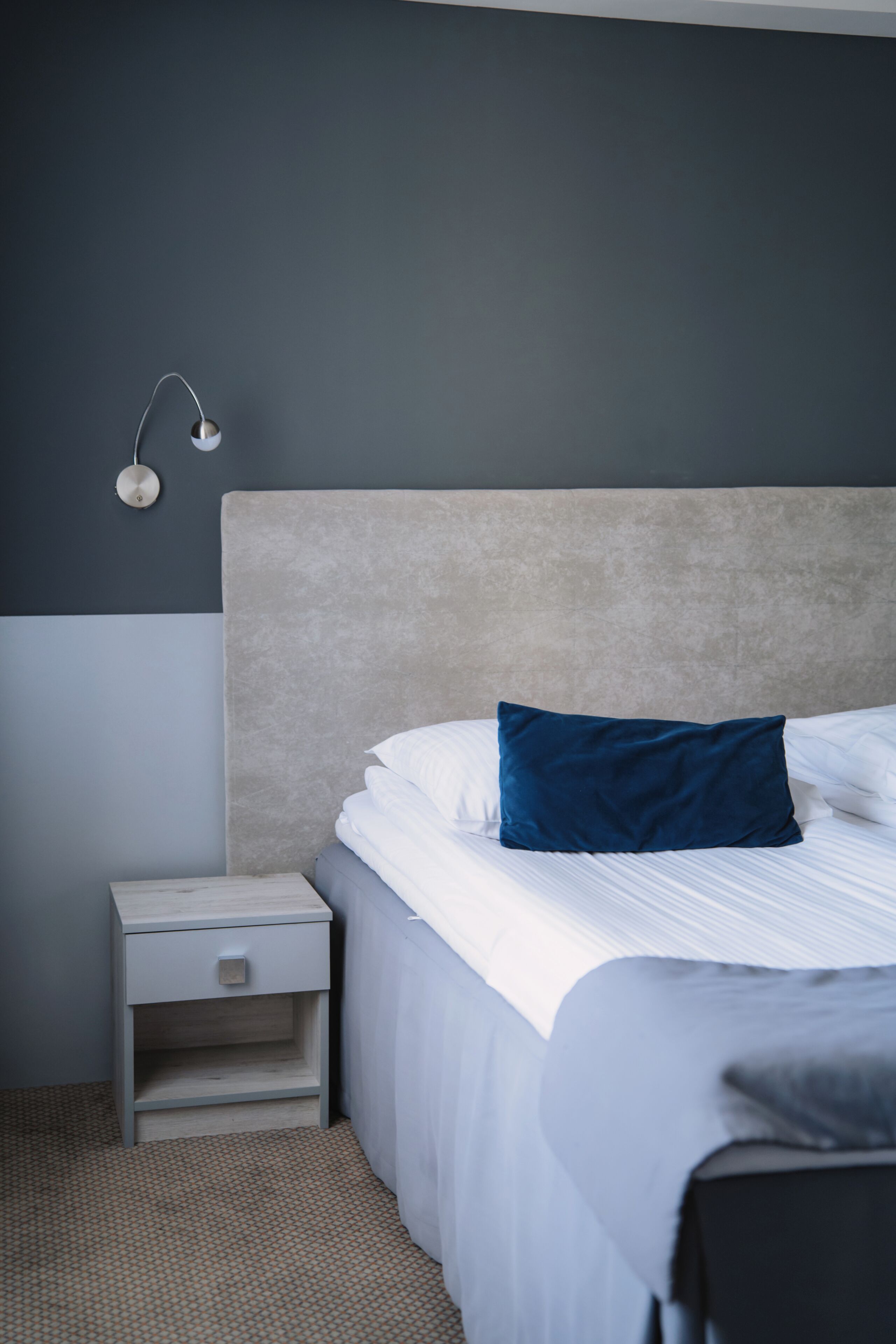 A bedroom with a bed, a nightstand, a wall-mounted lamp, and a patterned carpet.