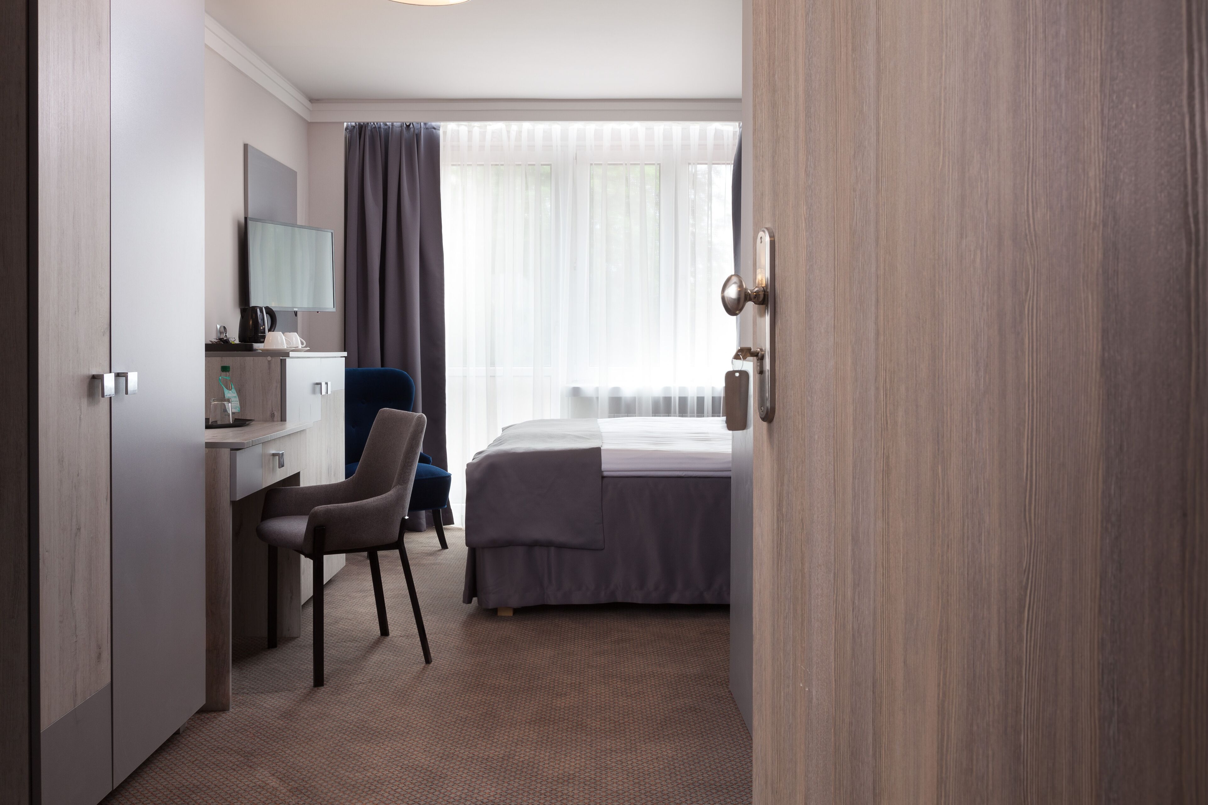 A hotel room with a bed, a desk with a chair, a television, and a window with curtains.
