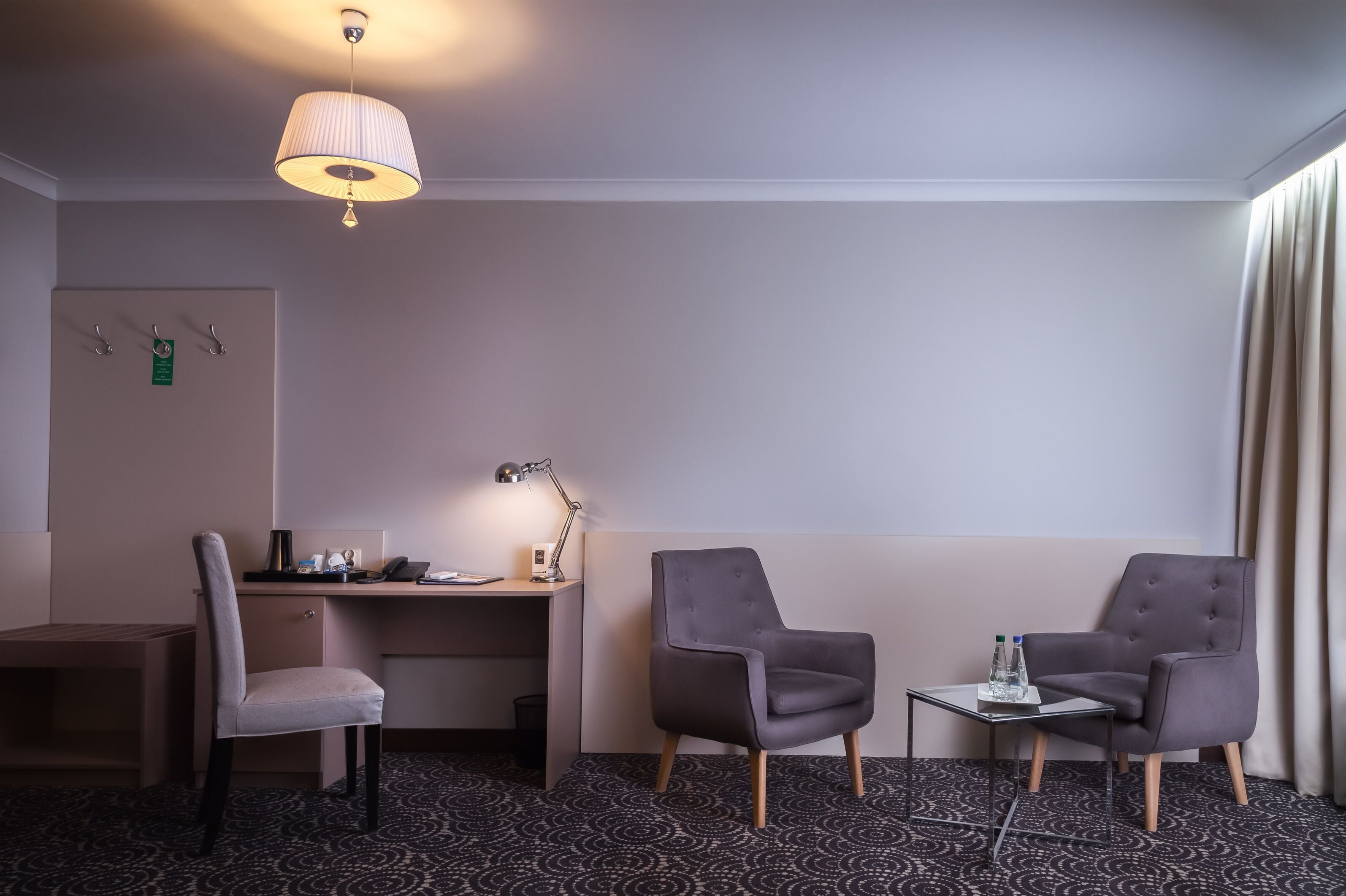 A hotel room with a desk, two chairs, a lamp, a small table with a bottle, and a patterned carpet.