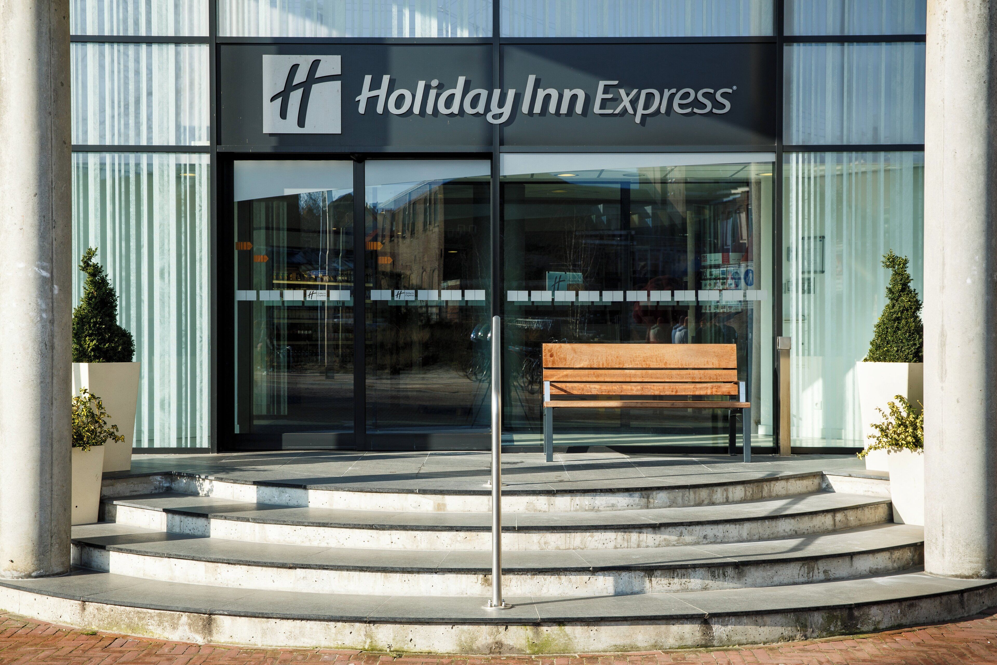The exterior of a Holiday Inn Express hotel with a bench in front, steps leading up to the entrance, and two potted plants on either side.