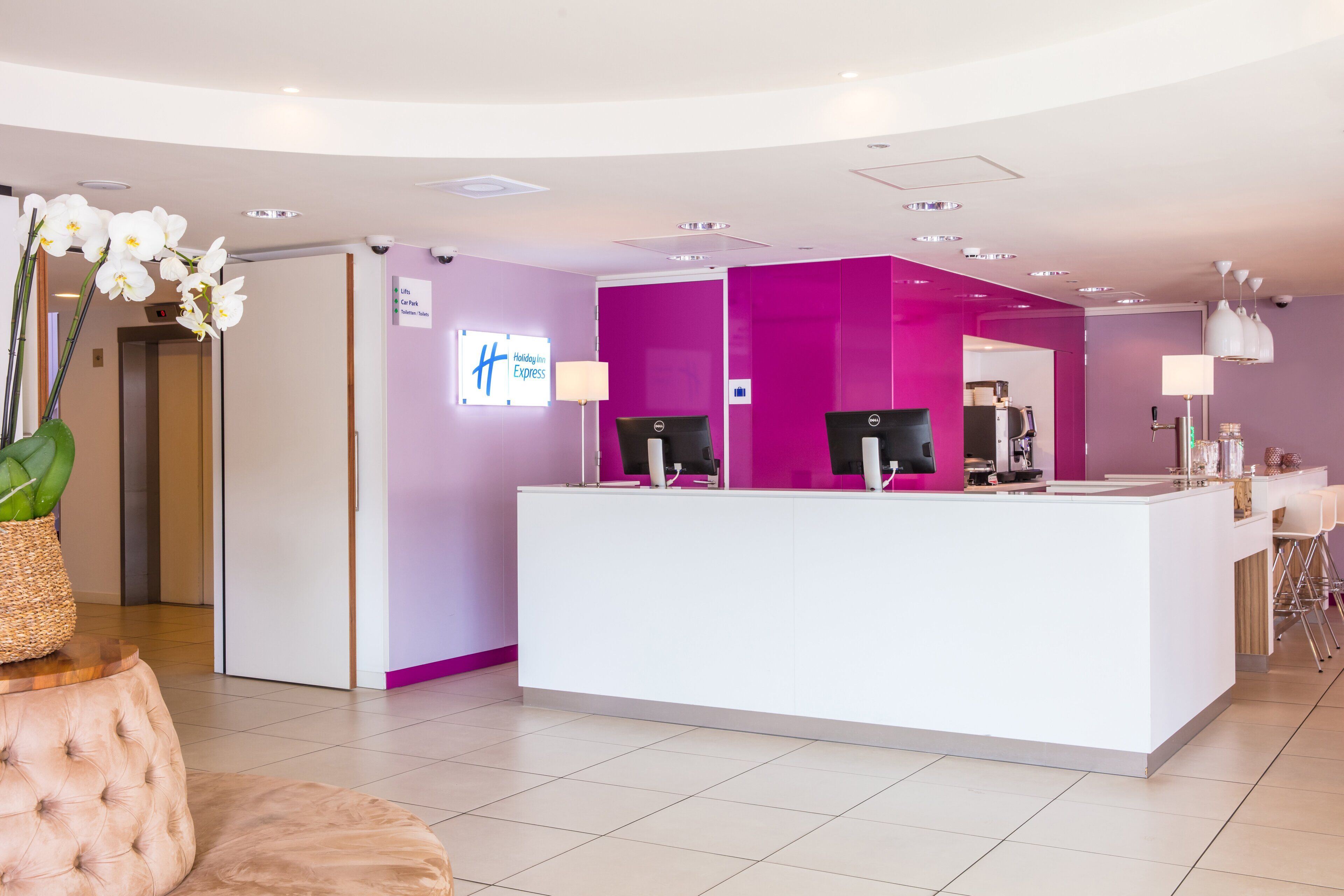 A hotel reception area with a white reception desk, purple accent walls, and a sign indicating amenities like lifts, car park, and toilets.
