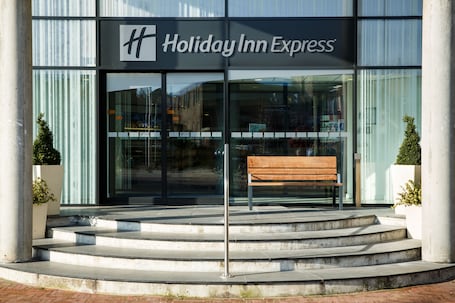 A Holiday Inn Express building with a modern glass facade, a wooden bench outside, and steps leading to the entrance.