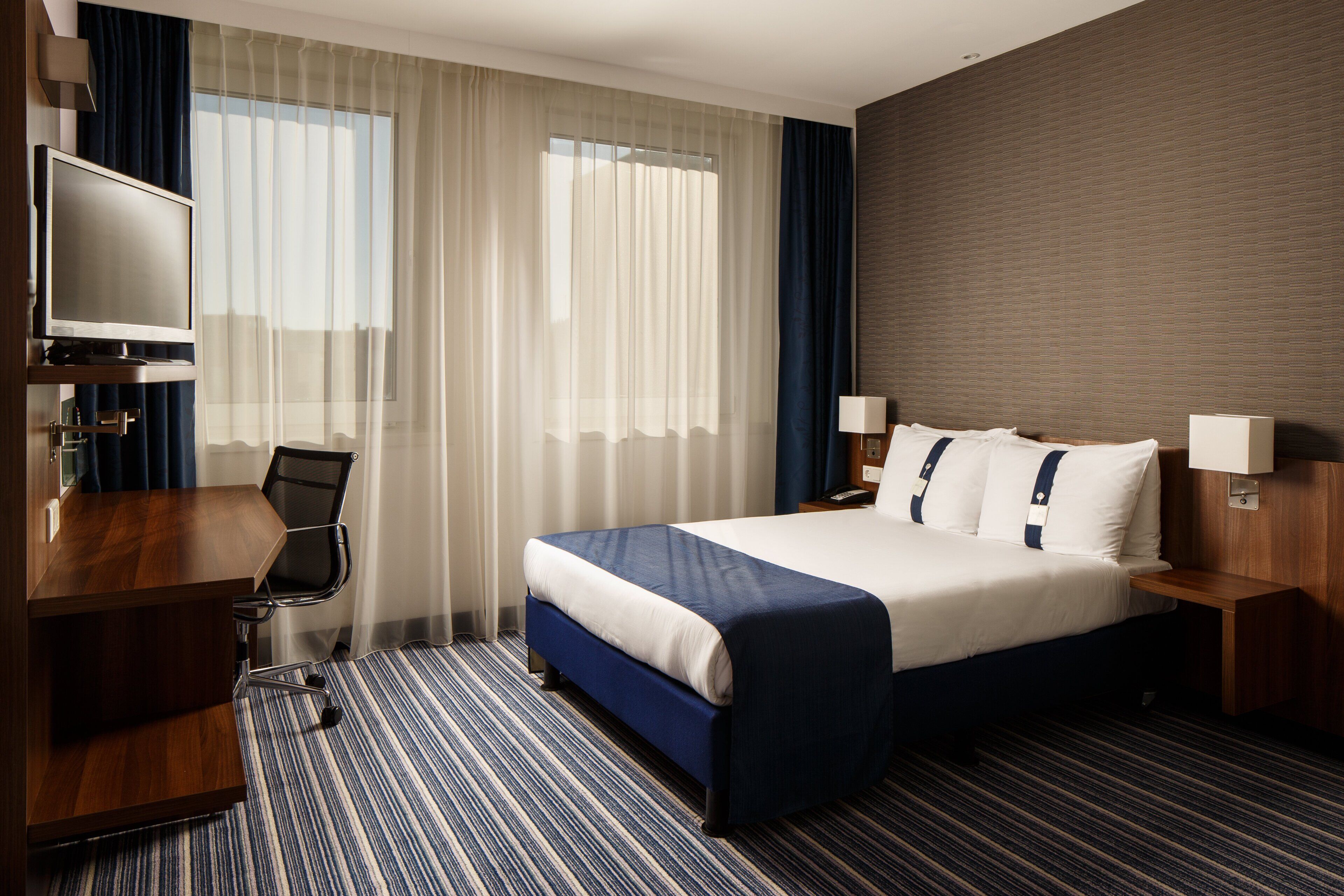 A hotel room with a bed, a desk, a chair, a TV, and a window with curtains.