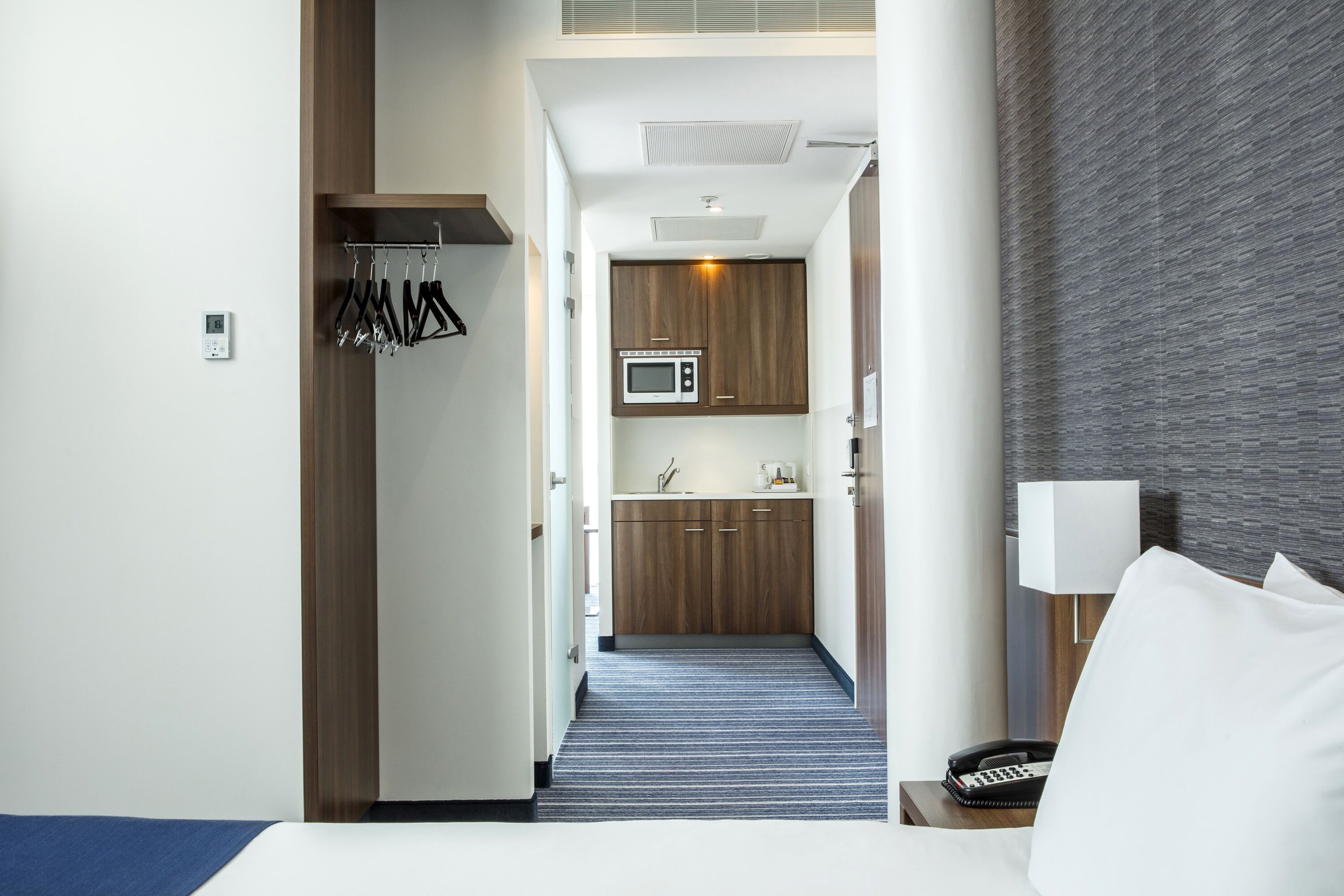 Een hotelkamer met een bed, een nachtkastje met een lamp, een kleine kitchenette met een magnetron en een badkamer door een geopende deur zichtbaar.