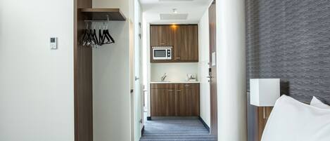 A hotel room with a bed, a bedside table with a lamp, a small kitchenette with a microwave, and a bathroom visible through an open door.