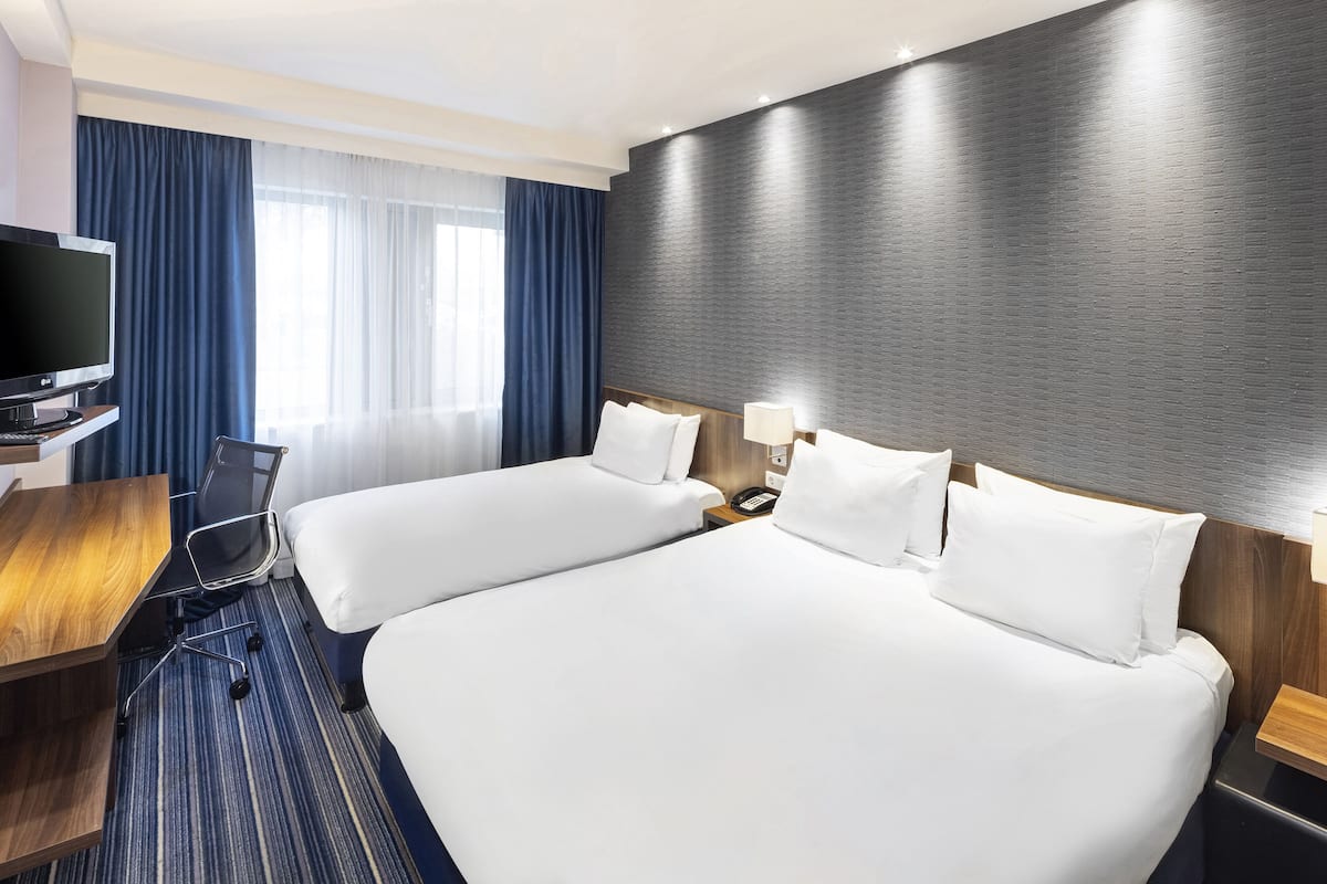 A hotel room with two beds, a desk, a chair, a TV, and a large window with curtains.