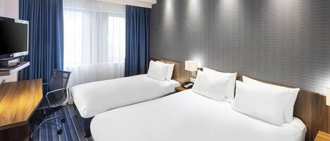 A hotel room with two beds, a desk, a chair, a TV, and a large window with curtains.