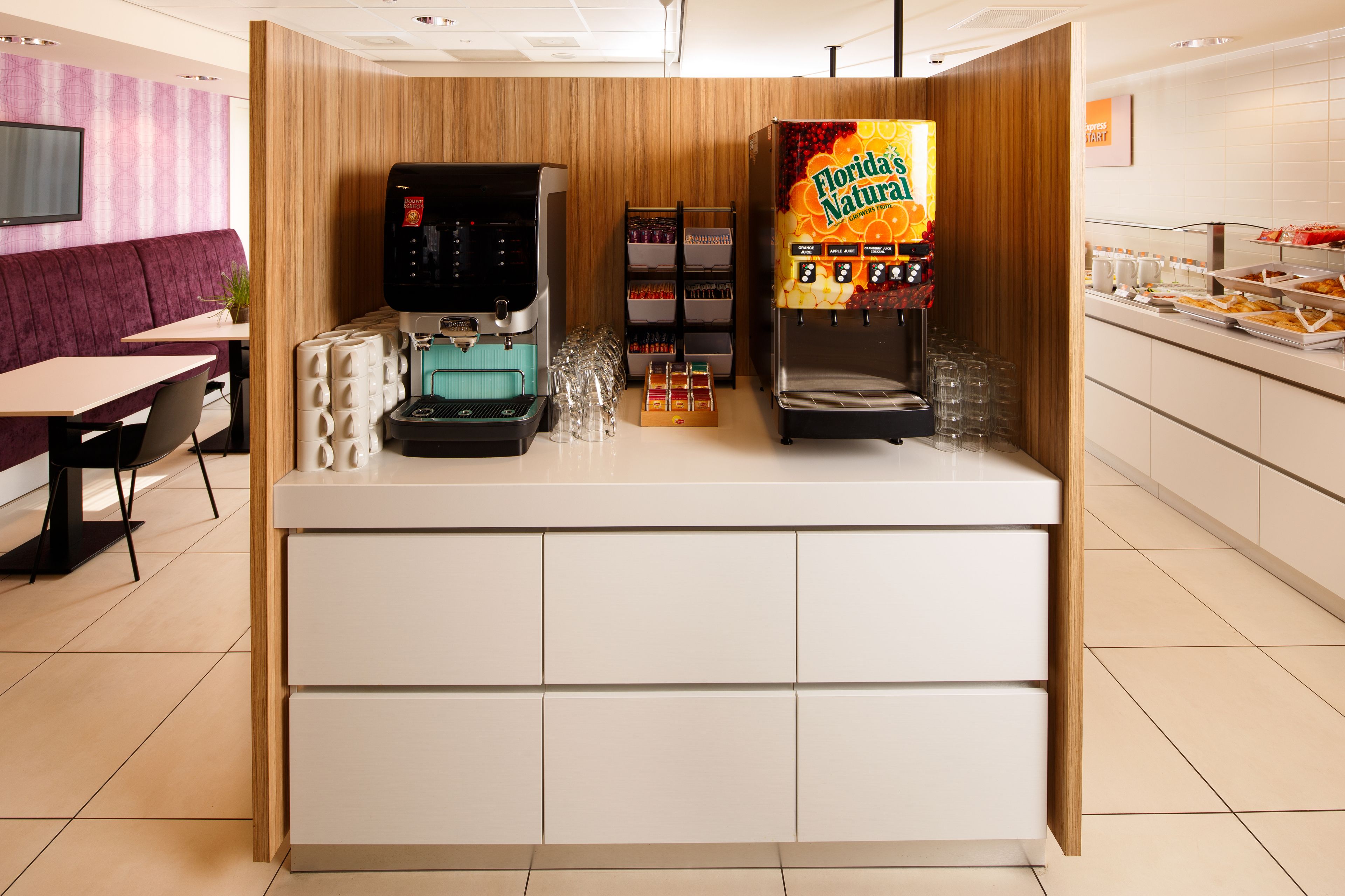 A coffee station with a coffee machine and a juice dispenser, a seating area with a purple booth, and a buffet counter with various food items.