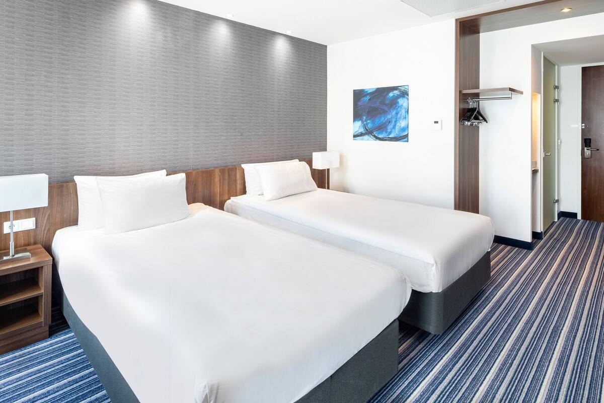 A hotel room with two beds, a nightstand with a lamp, a coat rack, and a door to the bathroom.