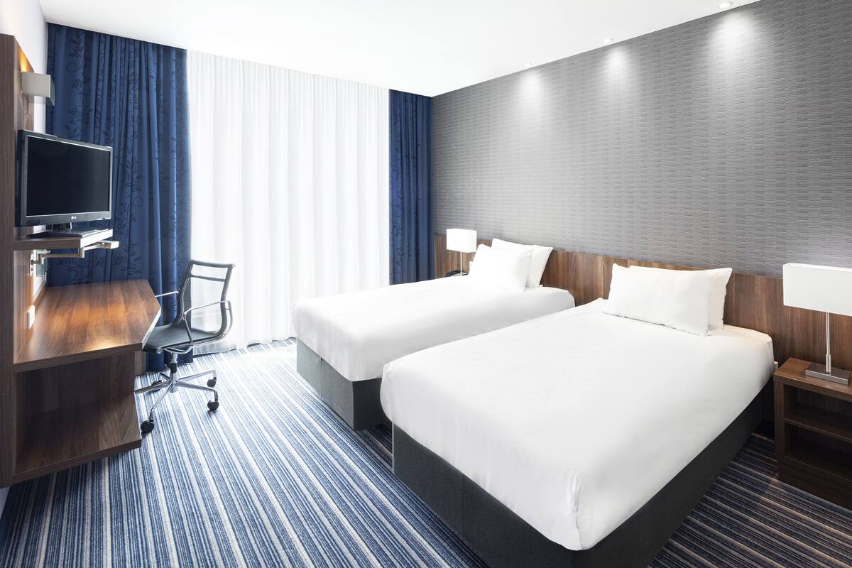 A hotel room with two beds, a desk with a TV, a chair, and a large window with curtains.