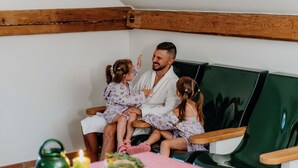 Eine dreiköpfige Familie in einem Zimmer mit Holzbalkendecke, einem grünen Sofa und einem Tisch mit Speisen und Getränken.