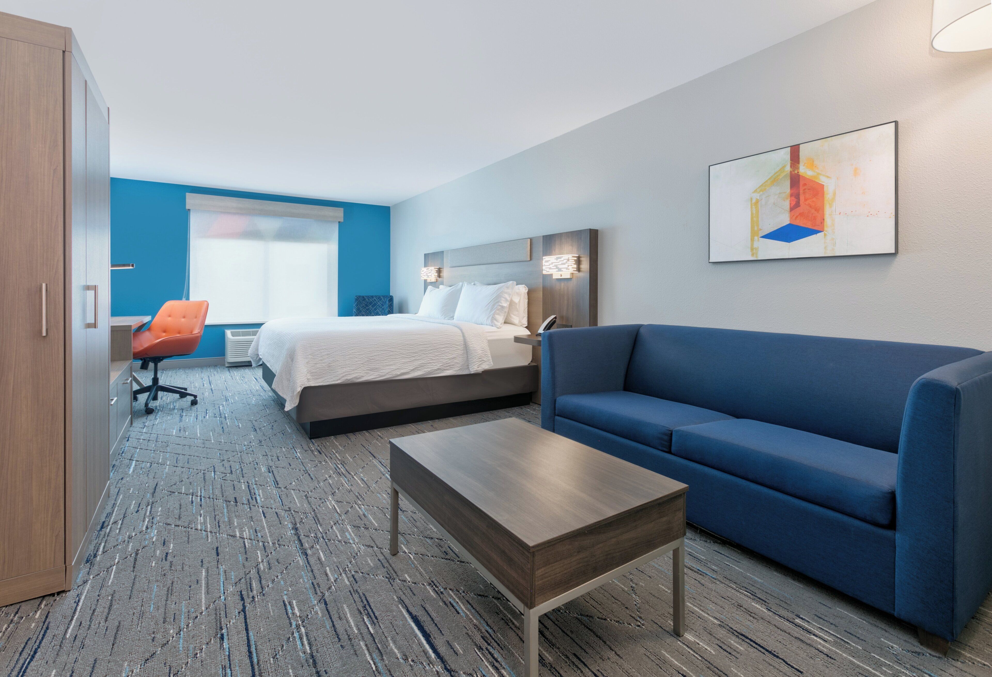 A hotel room with a bed, a blue sofa, a wooden coffee table, and a desk with an orange chair.