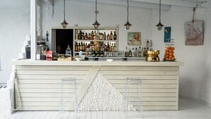 A bar area with a white wooden counter, hanging lights, and various bottles and glasses. - Ariadni Blue Hotel (Sithonia)