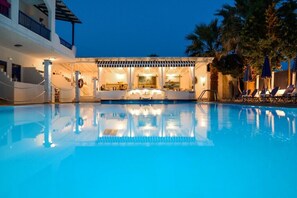 A pool area with a bar and lounge chairs, surrounded by palm trees and a building with multiple balconies. - Ariadni Blue Hotel (Sithonia)