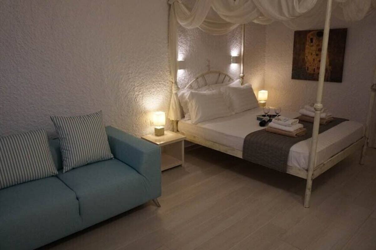 A bedroom with a four-poster bed, a nightstand with a lamp, a blue sofa with striped pillows, and a painting on the wall.
