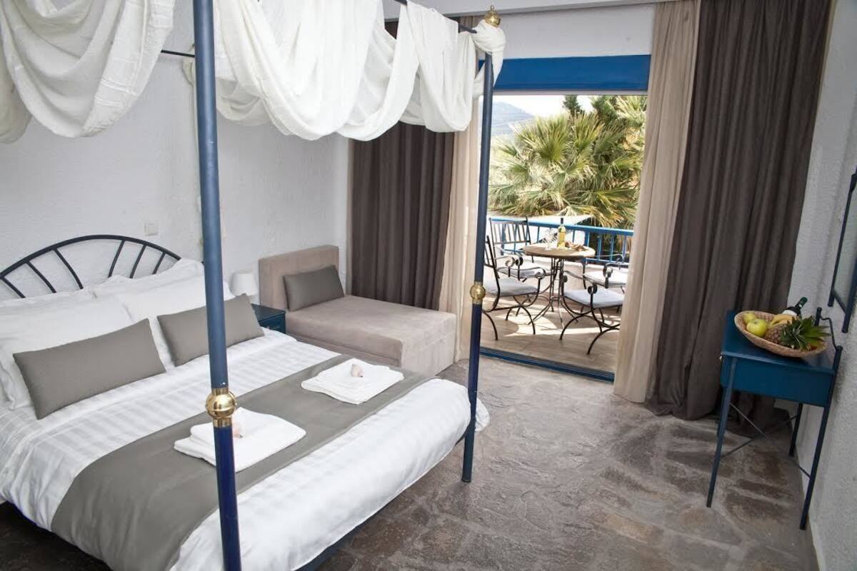 A bedroom with a four-poster bed, a sofa, a small table with fruit, and a balcony with a table and chairs.