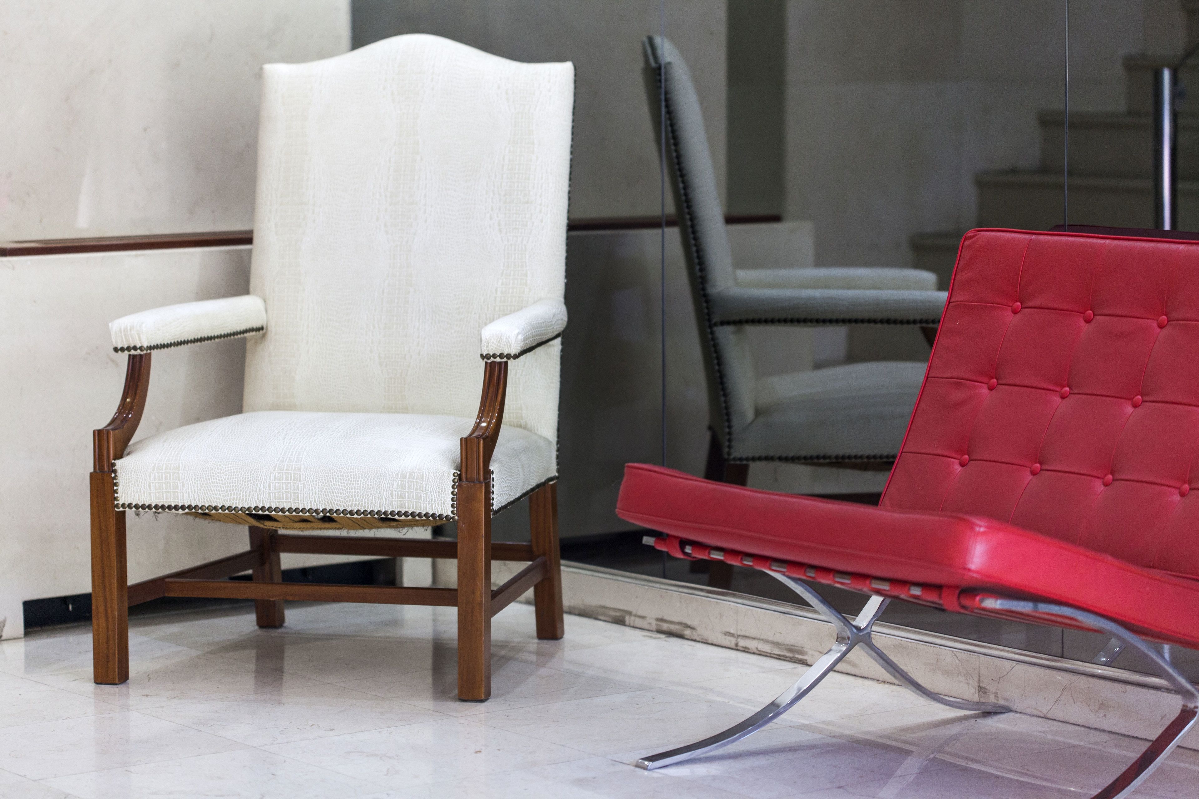 A chair with a wooden frame and white fabric seat, a red upholstered chair, and a grey upholstered chair in a room with a tiled floor.