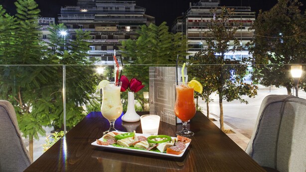 A table with a variety of food and drinks, a menu, and a view of buildings at night. - Leto Boutique Hotel (Agrinio)