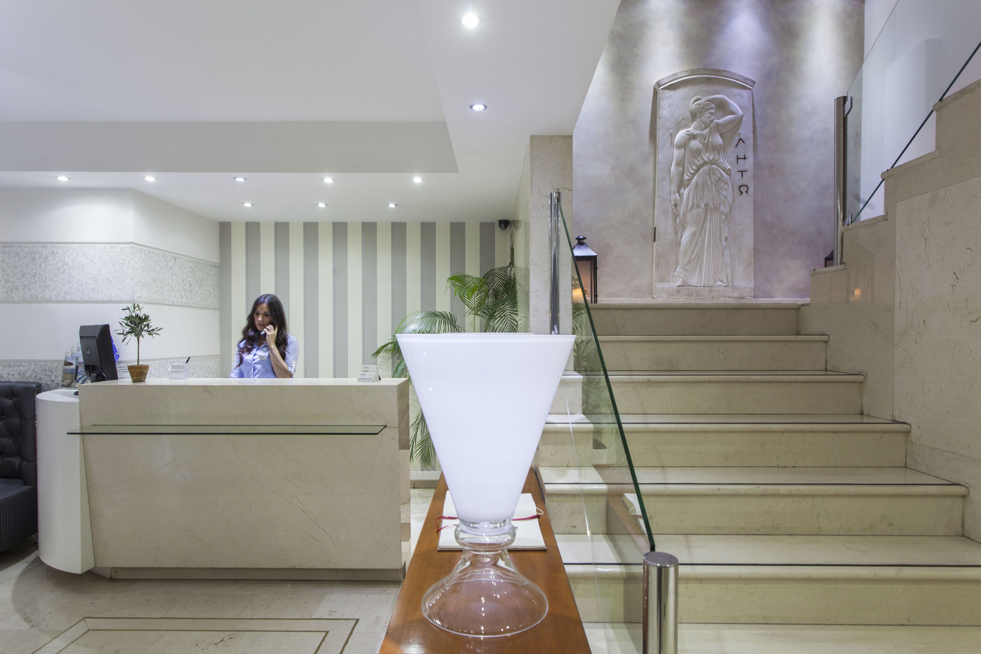 A reception area with a receptionist, a reception desk, a large white vase, a staircase, and a statue.