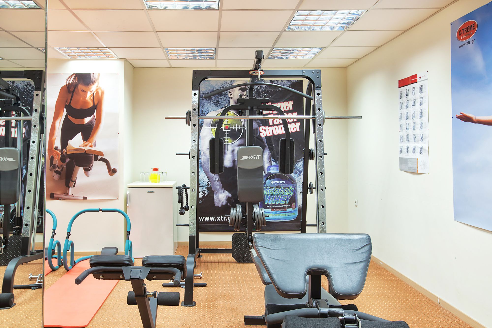 Uma academia com equipamentos de ginástica, um cartaz de uma mulher se exercitando e uma parede com um calendário.