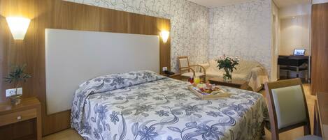 A hotel room with a large bed, a desk, a chair, a nightstand, and a potted plant.