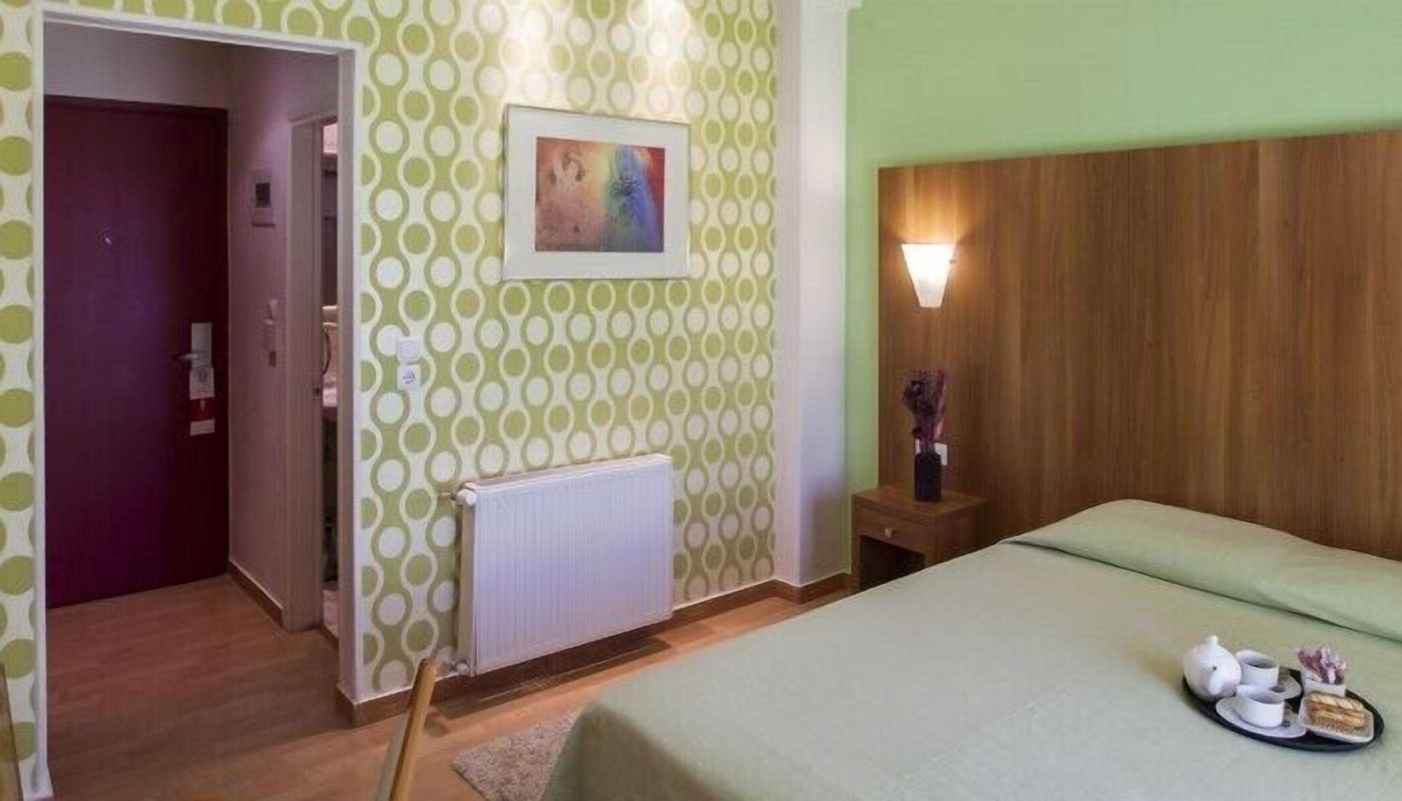 Quarto de hotel com cama, mesa de cabeceira com luminária, quadro na parede e porta para outro cômodo.