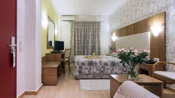 Habitación de hotel compacta con cama, escritorio, silla, mesita pequeña, mesita de noche, espejo y un jarrón con flores.