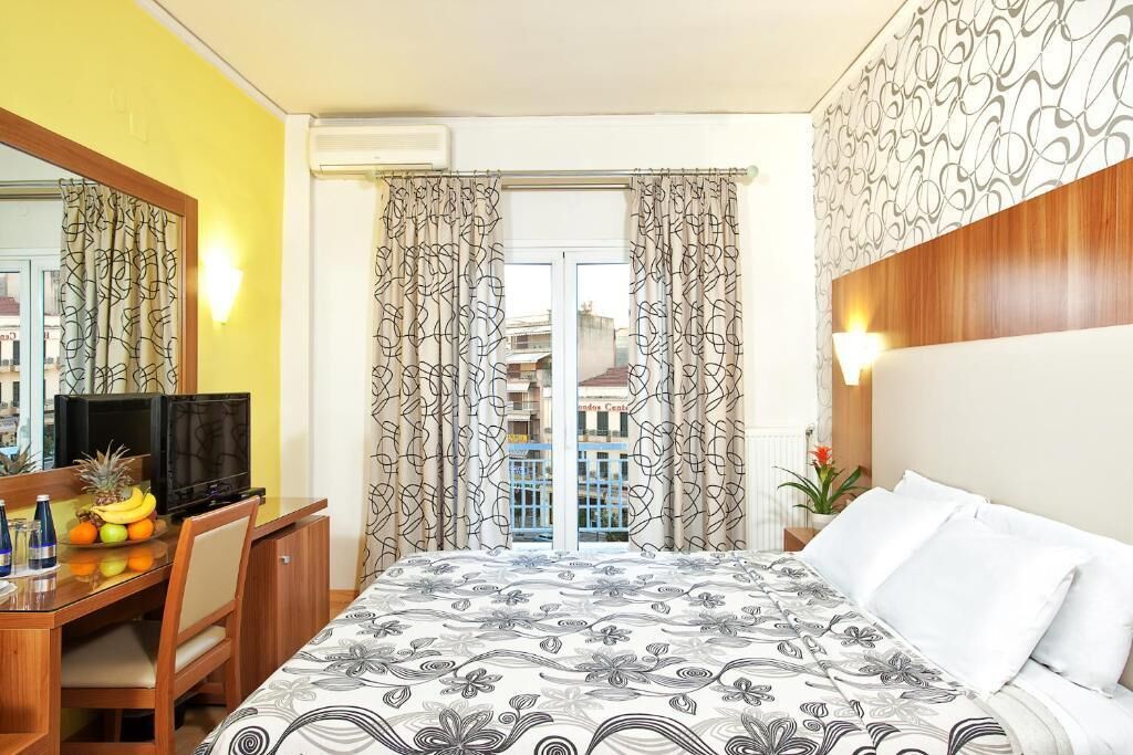 A hotel room with a bed, a desk with a TV, and a view of buildings through the window.