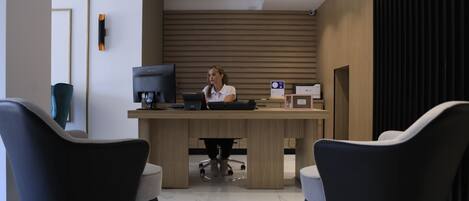 A modern reception desk with a woman seated behind it, two chairs in front, and a wooden panel wall.