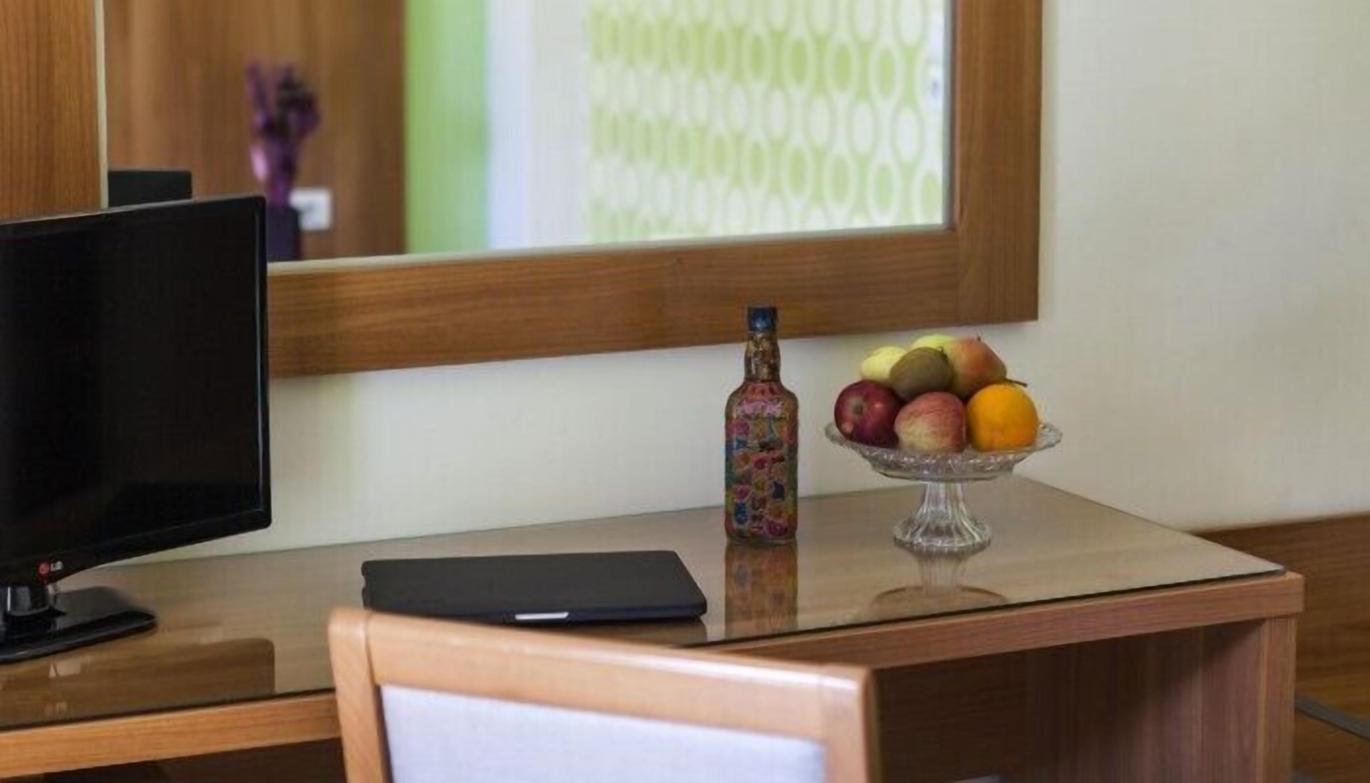 A hotel room with a wooden desk, a black monitor, a glass fruit bowl with apples and oranges, and a decorative bottle.