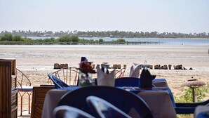 Vista frente a la playa con zona de comedor con sillas y mesas de metal, una playa arenosa y una costa lejana con árboles y agua.