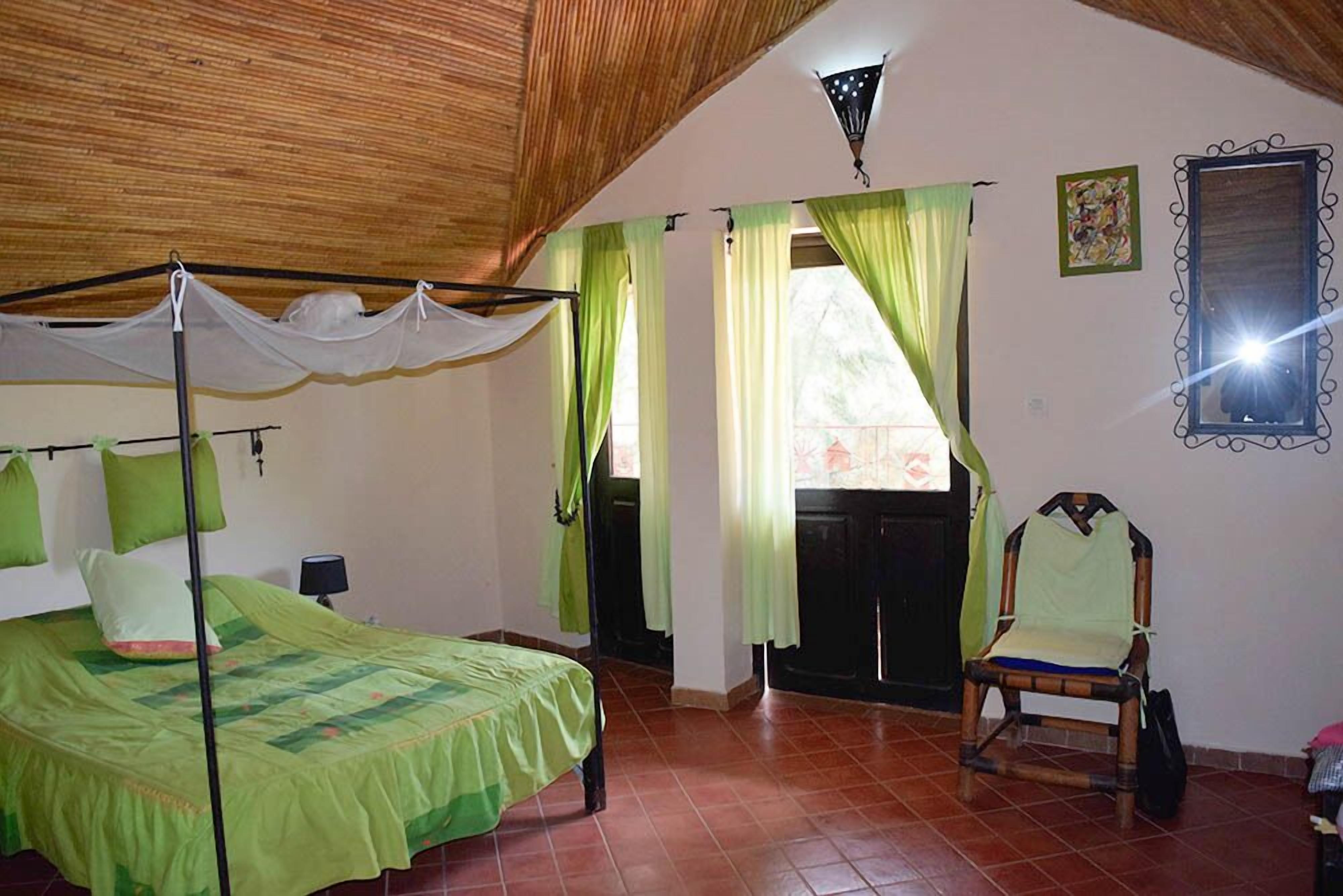 A bedroom with a bed, a chair, a window with curtains, a mirror, and a wooden ceiling.