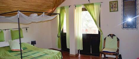 A bedroom with a bed, a chair, a window with curtains, a mirror, and a wooden ceiling.