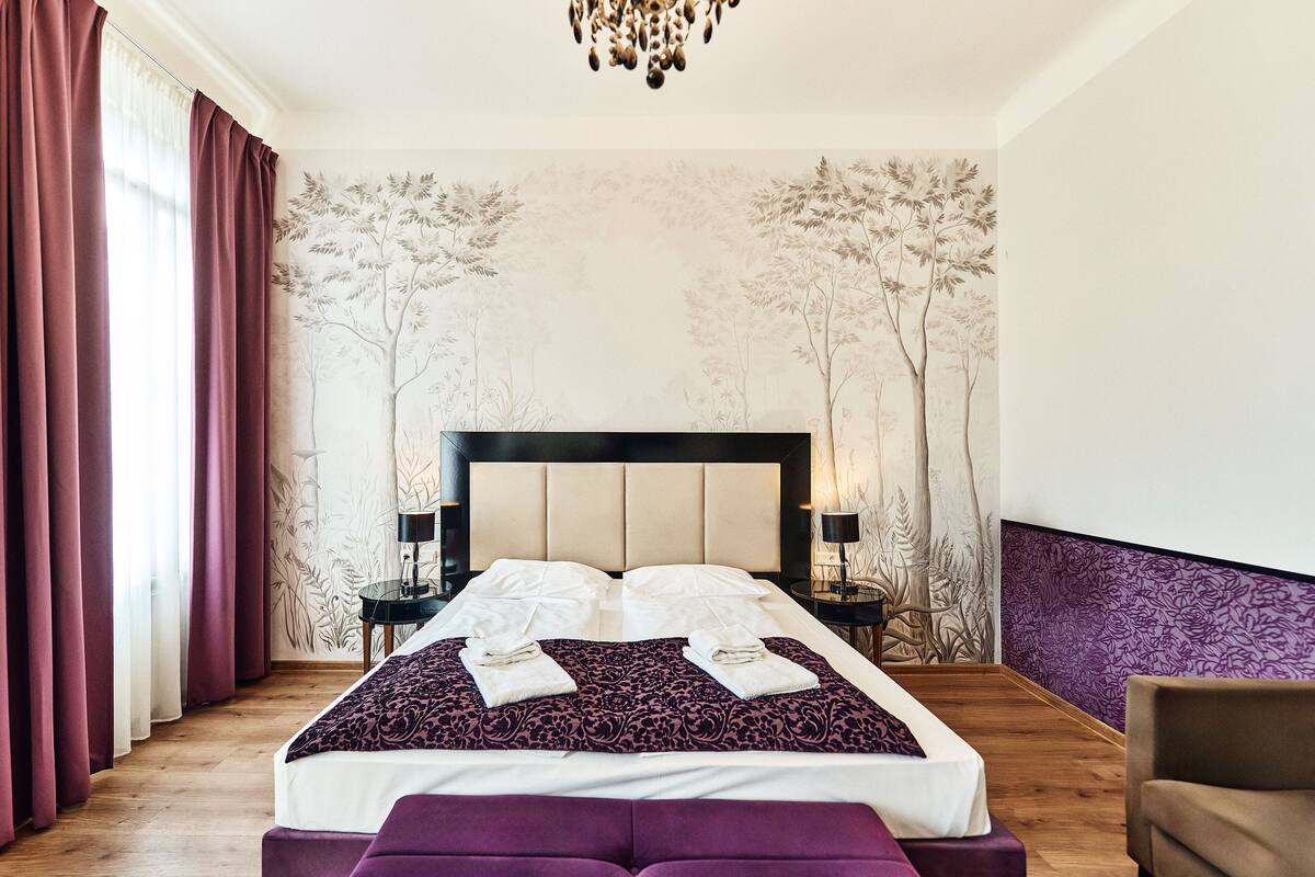 Una habitación de hotel con una cama grande, un cabecero, dos mesitas de noche con lámparas, un banco morado y una pared con un mural de un árbol.