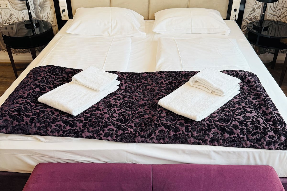 Una cama bien hecha con sábanas blancas y una colcha de flores moradas, un banco morado a juego, y dos mesitas de noche negras con lámparas.