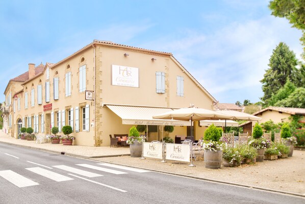 A two-story building with a sign reading "Hotel Restaurant Commerce." There are outdoor tables and chairs under umbrellas, and potted plants are visible.