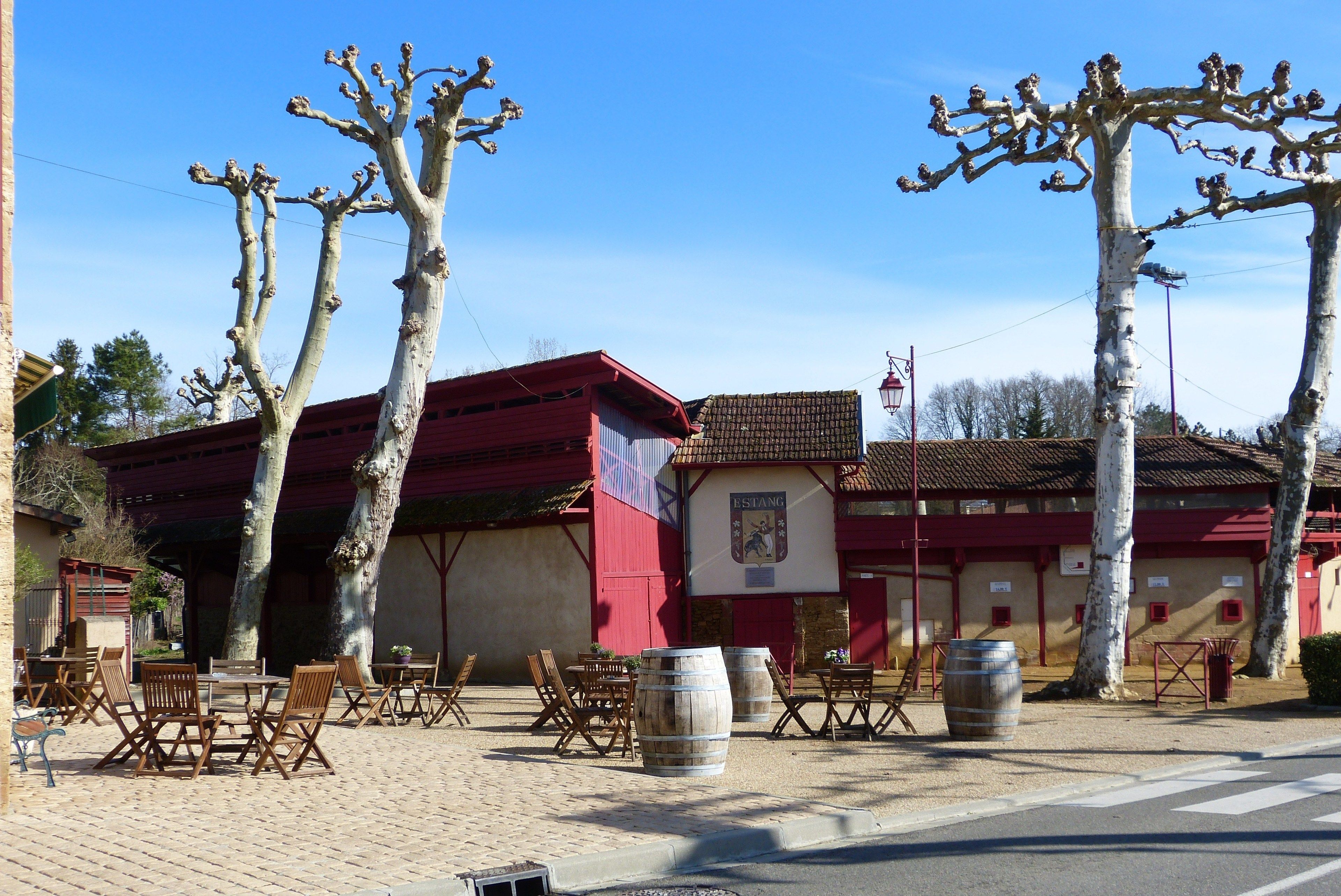 An outdoor seating area with tables and chairs, wooden barrels, and a building with a red roof.