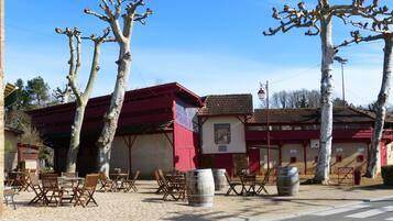 An outdoor seating area with tables and chairs, wooden barrels, and a building with a red roof.