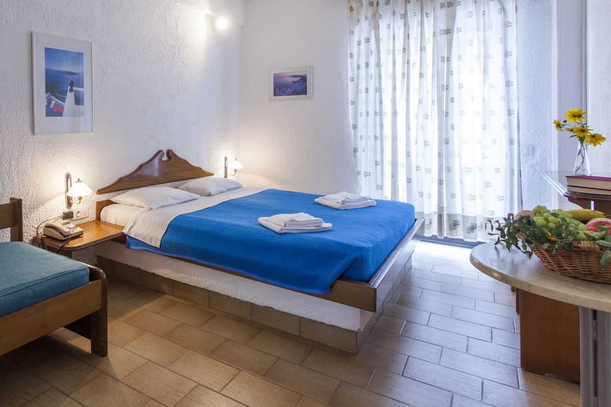 Une chambre à coucher avec un lit, une table de chevet avec une lampe, une chaise, une fenêtre avec des rideaux et une petite table sur laquelle se trouve un panier de fruits.