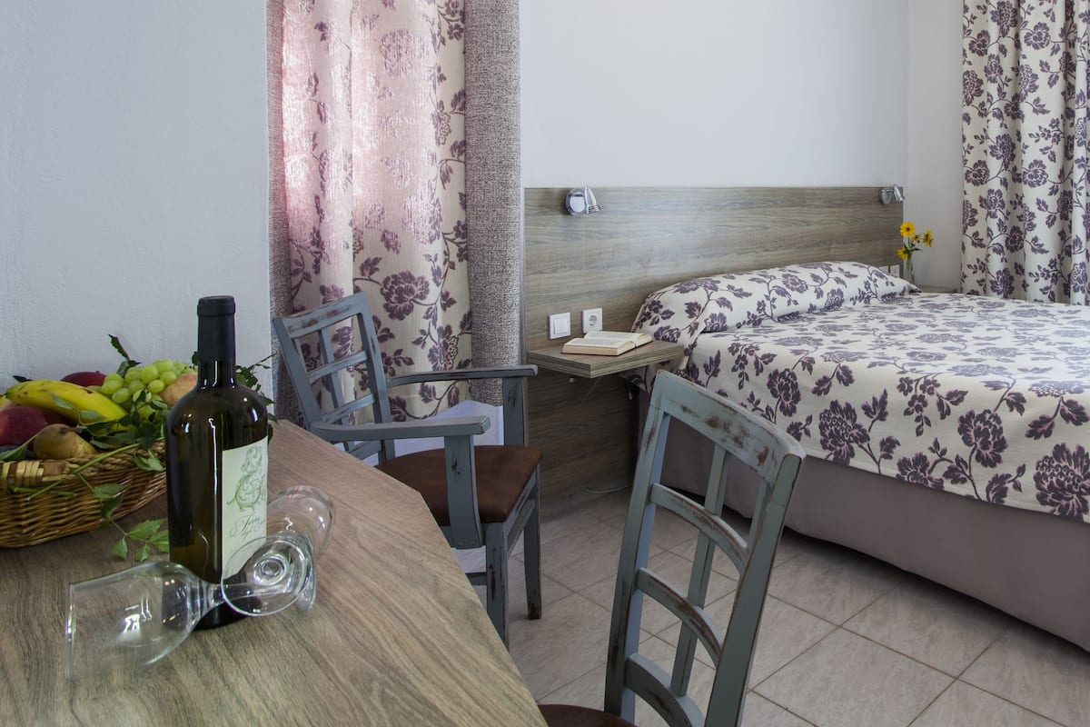Une chambre à coucher avec un lit, une table de chevet sur laquelle se trouvent un livre et un vase, une chaise, une table avec une bouteille et des verres, et une fenêtre avec des rideaux.
