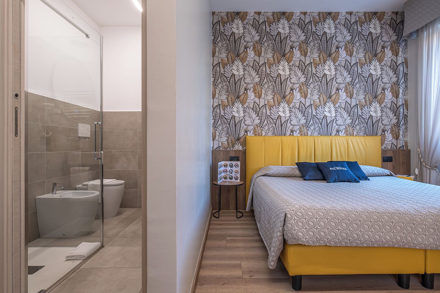 Camera d'albergo moderna con box doccia in vetro, un letto con testiera gialla e carta da parati decorata.