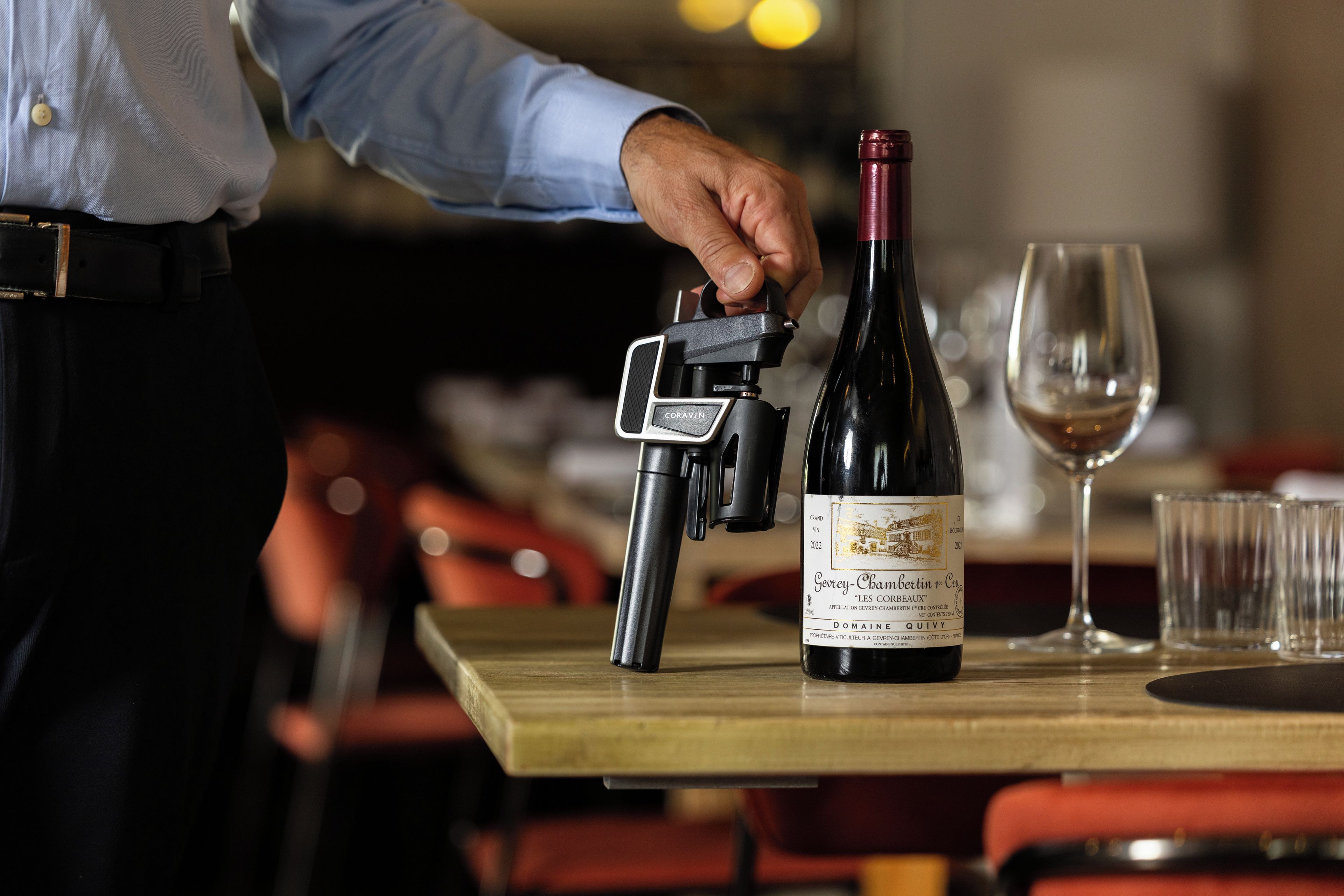Une bouteille de vin avec une étiquette posée sur une table, un verre à vin et une personne tenant un appareil photo.