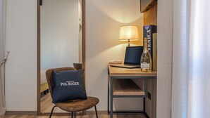 Een hotelkamer met een houten bureau, een laptop, een fles champagne, een lamp en een stoel met een kussen.