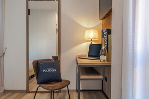 Habitación de hotel con un escritorio de madera, una computadora portátil, una botella de champán, una lámpara y una silla con un cojín.