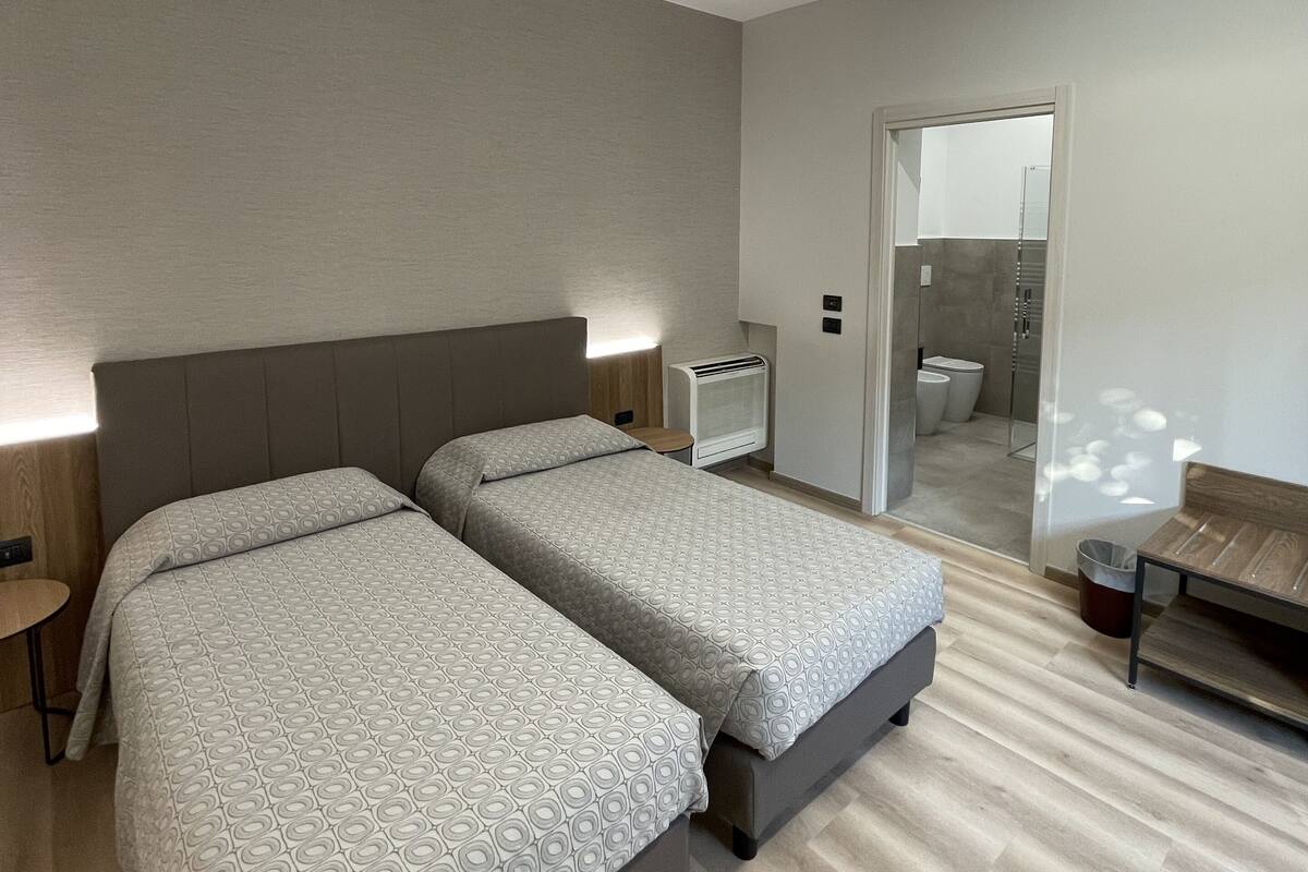 A modern hotel room with two beds, a small table, a bench, and a bathroom visible through a glass door.