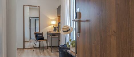 A hotel room with a wooden floor, a desk with a lamp, a chair, a suitcase, and a hat on the wall.
