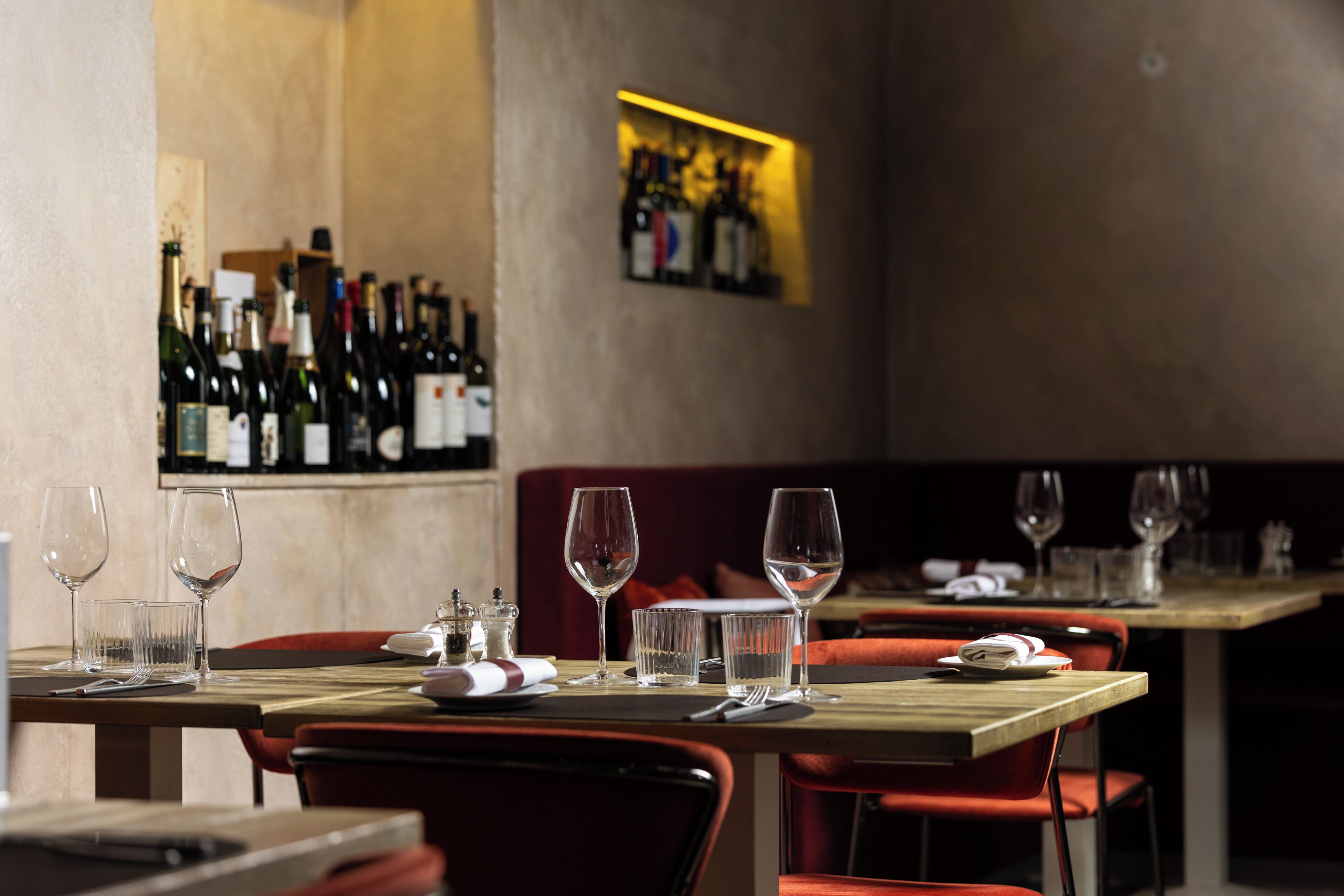 Un interior de restaurante con mesas dispuestas para comer, botellas de vino en una estantería y un cuadro en la pared.
